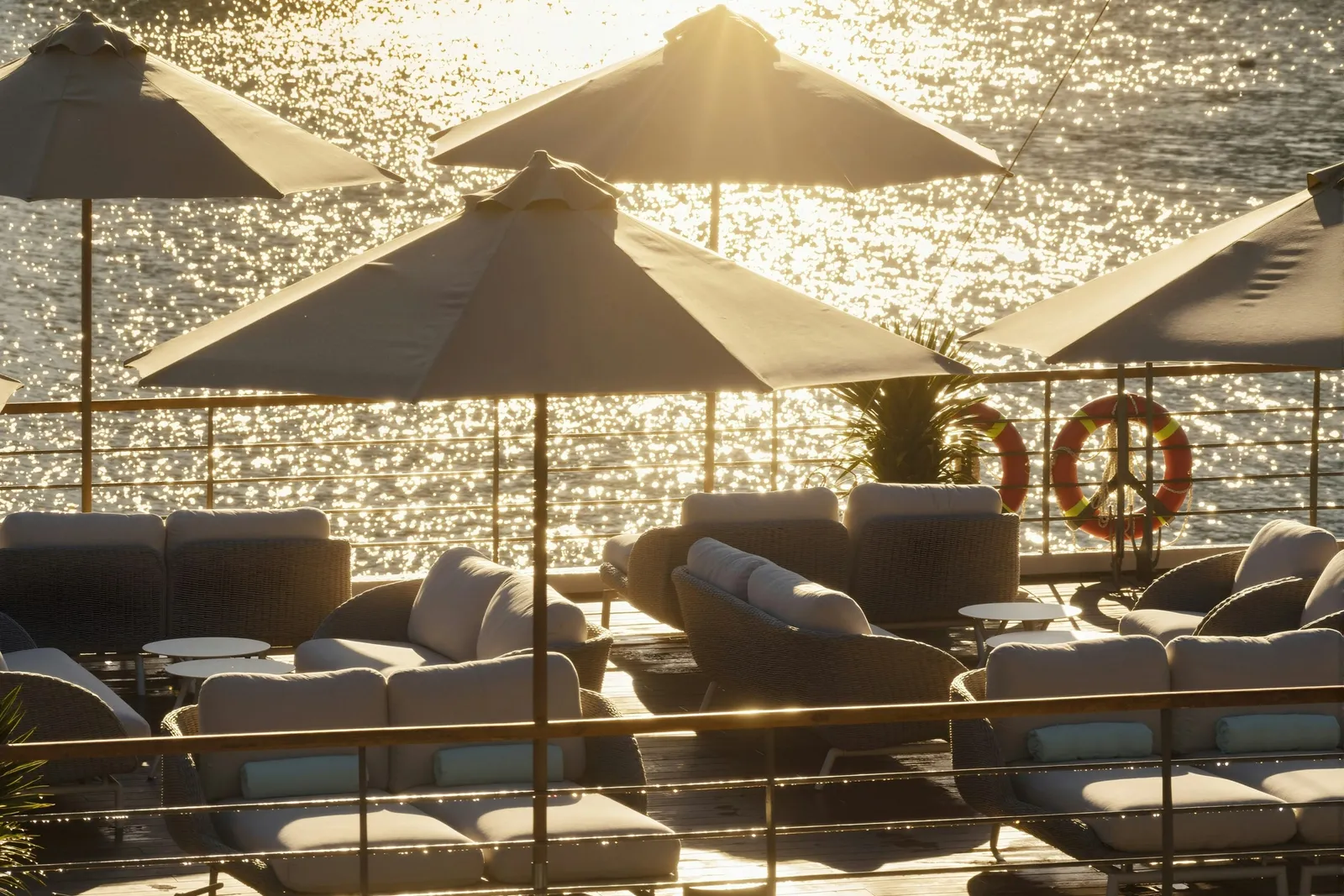 A boat's sundeck at golden hour with wicker lounge seating, cream parasols, orange life rings on the railing, and sunlit glittering water behind.