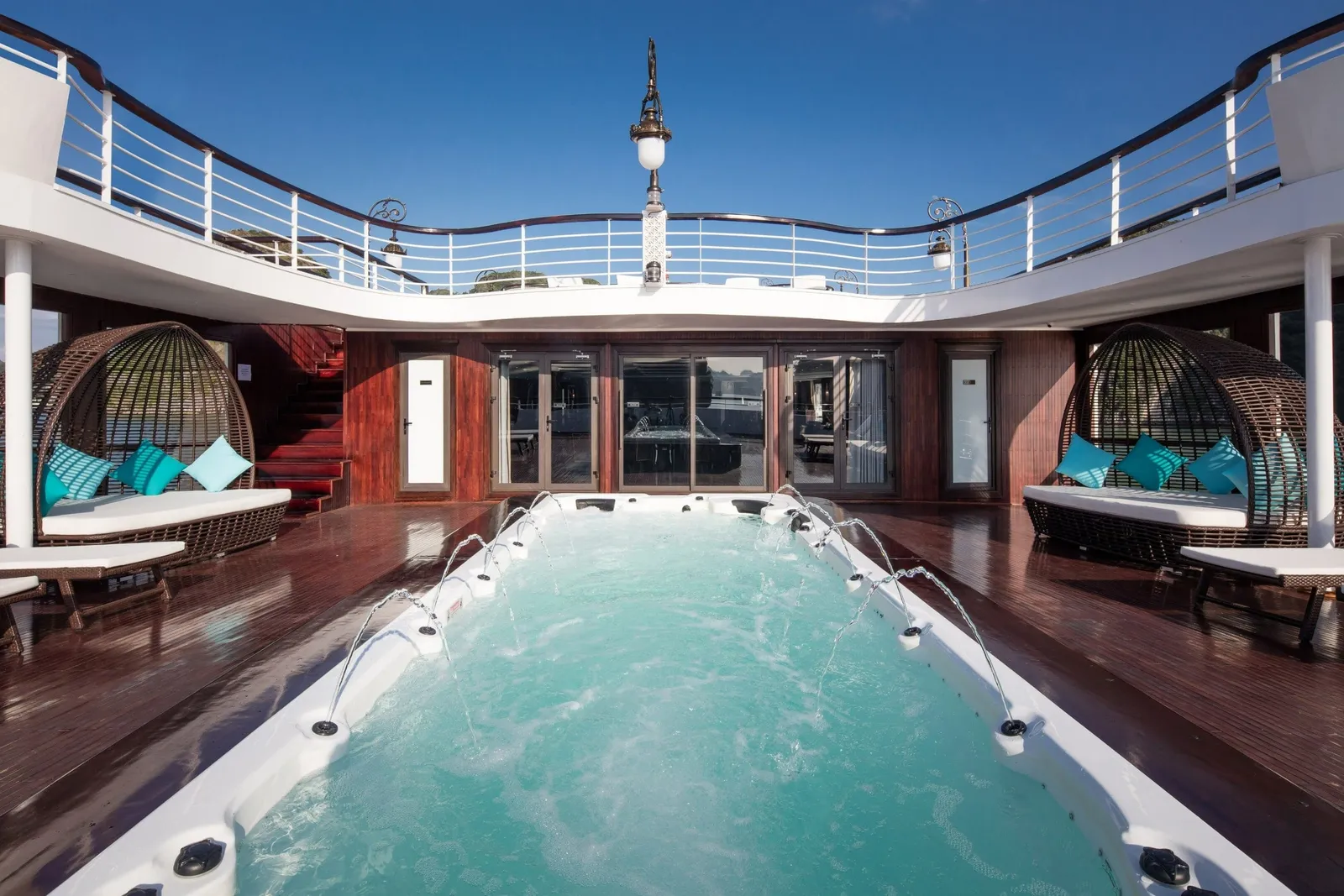 A wooden sun deck on a boat with a jetted plunge pool in the foreground, wicker daybeds with teal cushions on both sides, a curved upper-deck railing, and a clear blue sky.