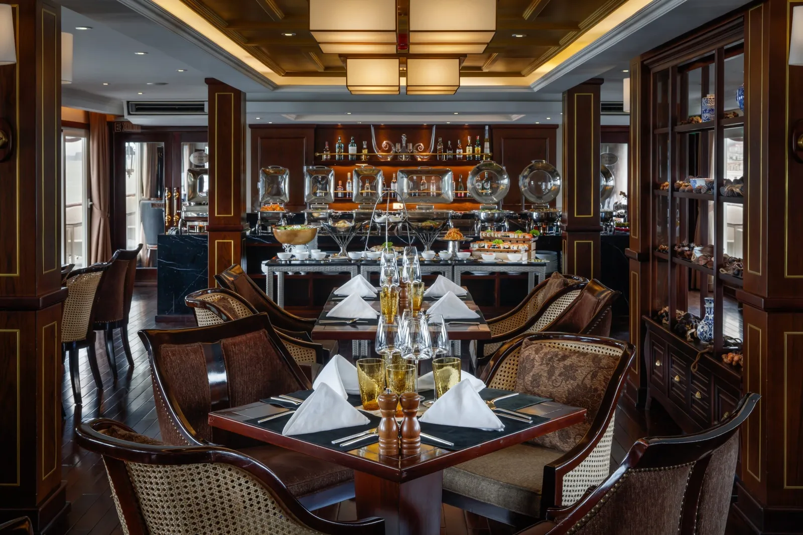 A dark-wood dining room with set tables, cane-back armchairs, folded white napkins and glassware, a buffet with chafing dishes at the back, and a display cabinet on the right.