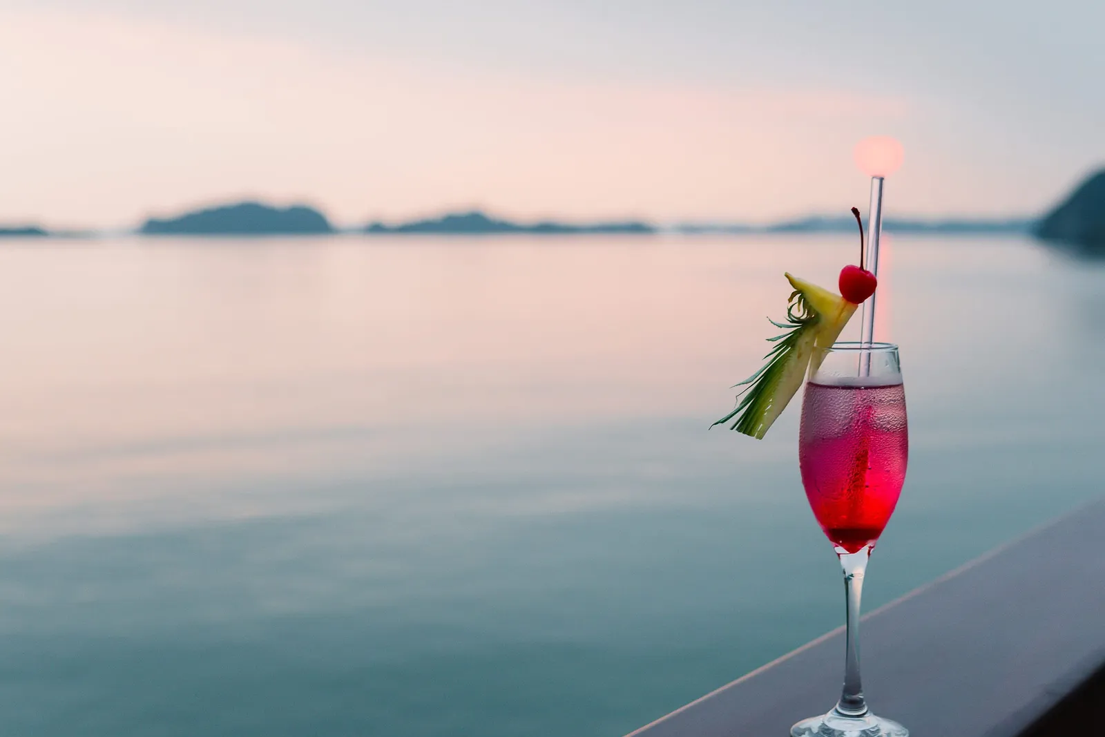 A pink cocktail garnished with pineapple and a cherry sits on a wooden boat railing in front of calm water and distant hazy islands at dusk.
