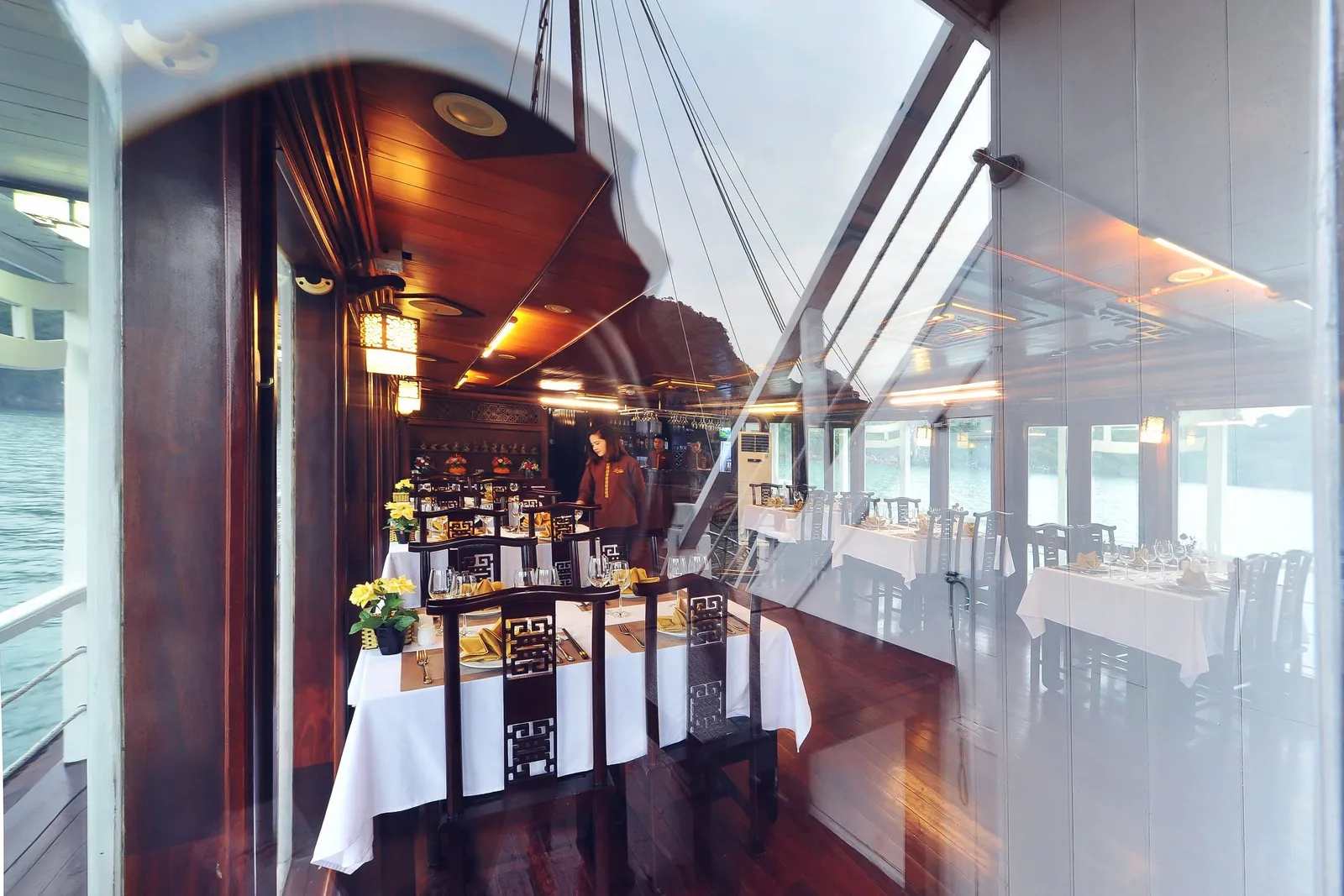 A set dining room on a boat with white-clothed tables, dark wooden chairs, yellow flowers and a staff member standing, photographed through a reflective glass window with water and a karst hill visible outside.