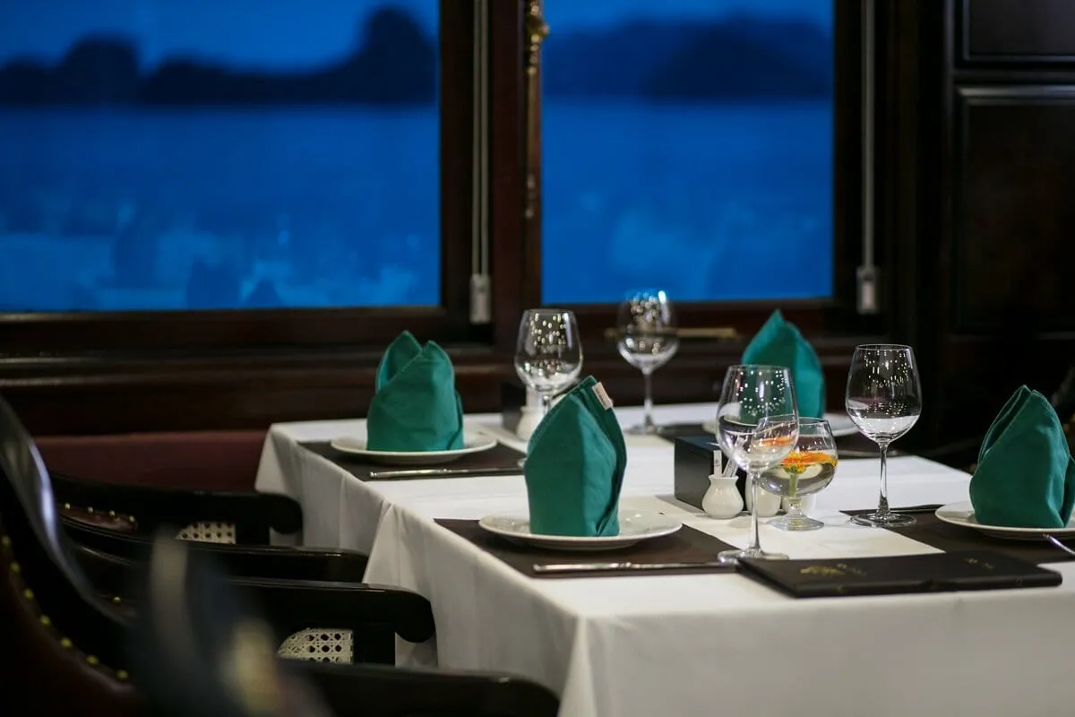 A dining table with white cloth, green folded napkins, wine glasses and menu folders sits beside a dark wood-framed window showing a blue bay with dark silhouetted islands.
