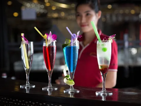 Four colorful cocktails with orchid garnishes and straws stand on a dark bar counter, with a smiling woman in a red polo shirt blurred behind them.