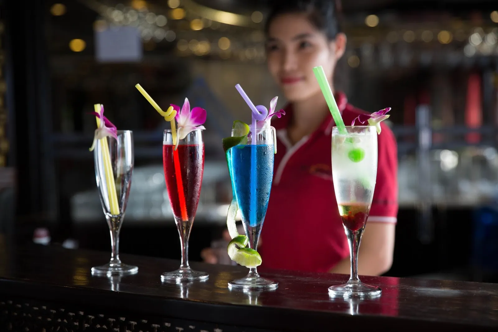 Four colorful cocktails with orchid garnishes and straws stand on a dark bar counter, with a smiling woman in a red polo shirt blurred behind them.