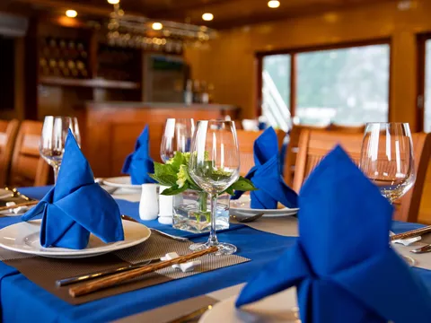 A wood-paneled dining room with tables set with blue tablecloths, folded blue napkins on white plates, wine glasses, chopsticks and a small vase of green leaves, with a bar behind and windows showing misty hills outside.