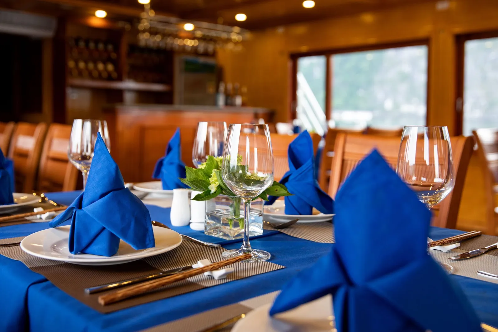 A wood-paneled dining room with tables set with blue tablecloths, folded blue napkins on white plates, wine glasses, chopsticks and a small vase of green leaves, with a bar behind and windows showing misty hills outside.
