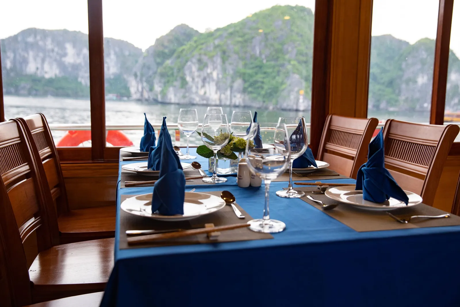 A dining table with a blue tablecloth, folded navy napkins, plates, wine glasses and chopsticks sits beside windows overlooking calm water and green limestone karst islands.