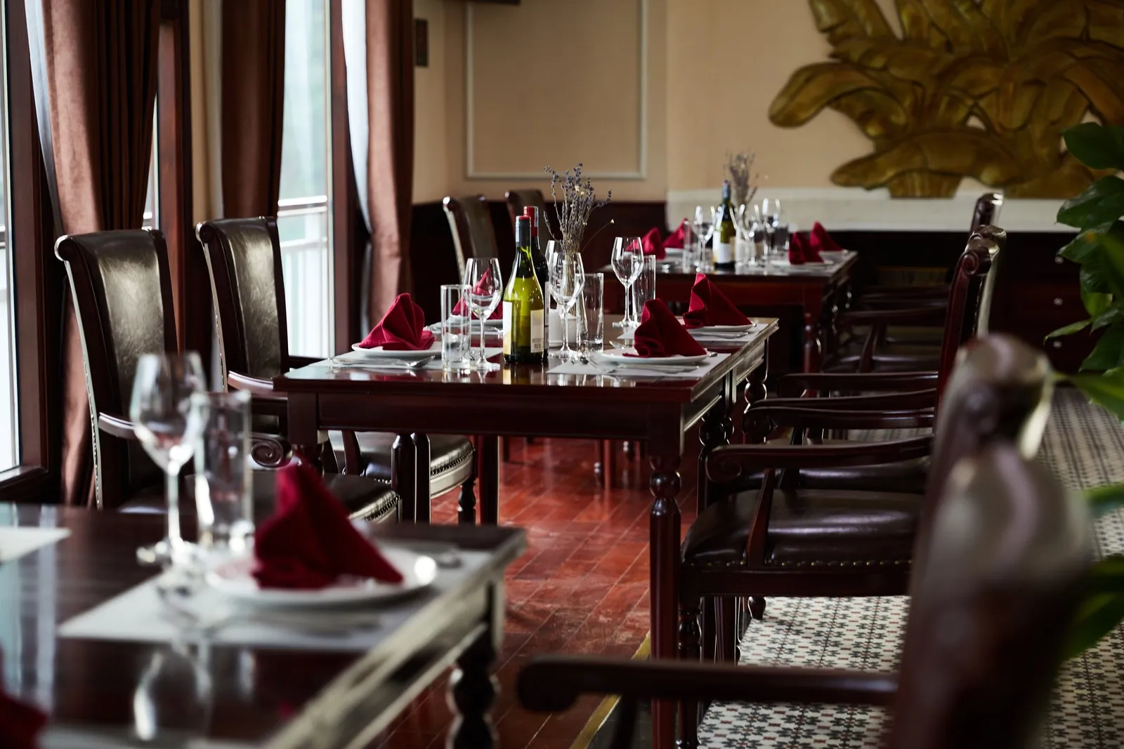 An indoor dining room with dark wood tables set with wine glasses, wine bottles and folded burgundy napkins, leather chairs, floor-length curtains, and a golden leaf wall decoration.