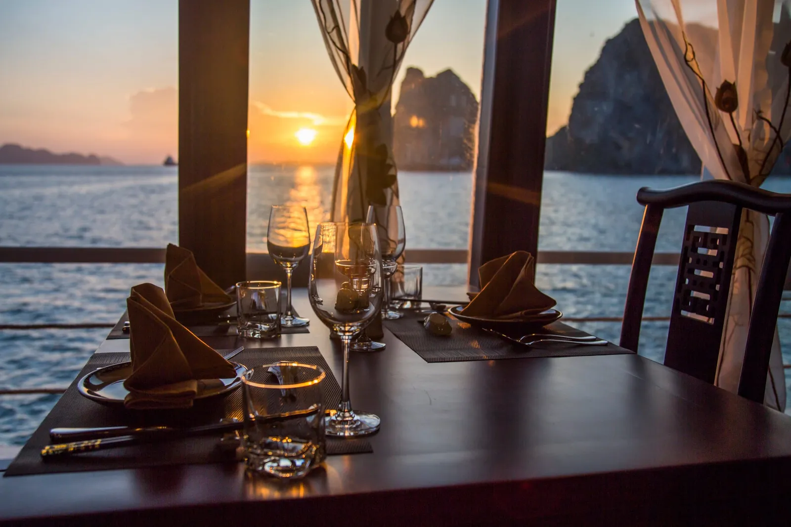 A dining table set with napkins, plates, and glasses sits beside open windows with curtains, looking out over the sea toward limestone karst rocks and a setting sun.