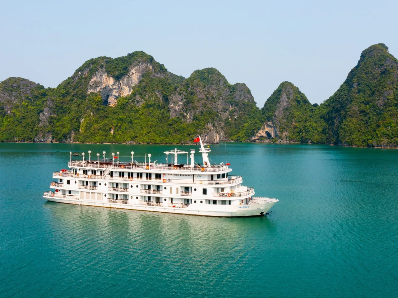 A multi-deck white cruise ship with small name lettering on its side sits on turquoise water in front of green limestone karst islands, photographed from an elevated vantage point off the vessel.