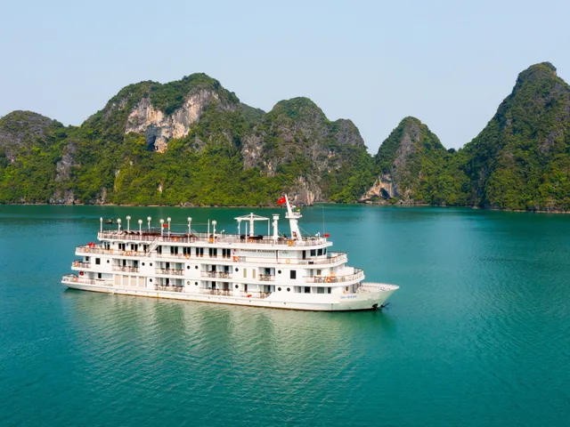A multi-deck white cruise ship with small name lettering on its side sits on turquoise water in front of green limestone karst islands, photographed from an elevated vantage point off the vessel.