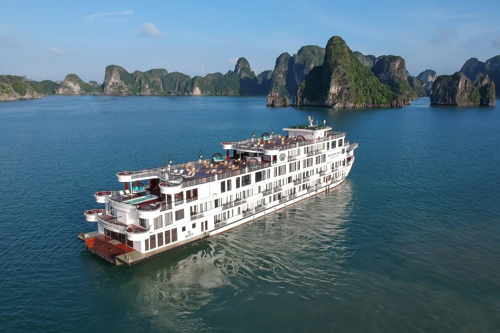 An aerial view of a large white multi-deck cruise ship with a sun deck, pool and a small logo and lettering on its hull, sailing on calm green water in front of tree-covered limestone karst islands.