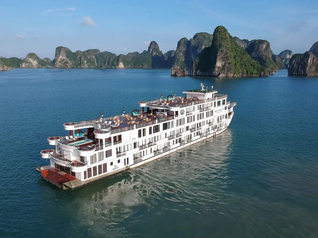 An aerial view of a large white multi-deck cruise ship with a sun deck, pool and a small logo and lettering on its hull, sailing on calm green water in front of tree-covered limestone karst islands.