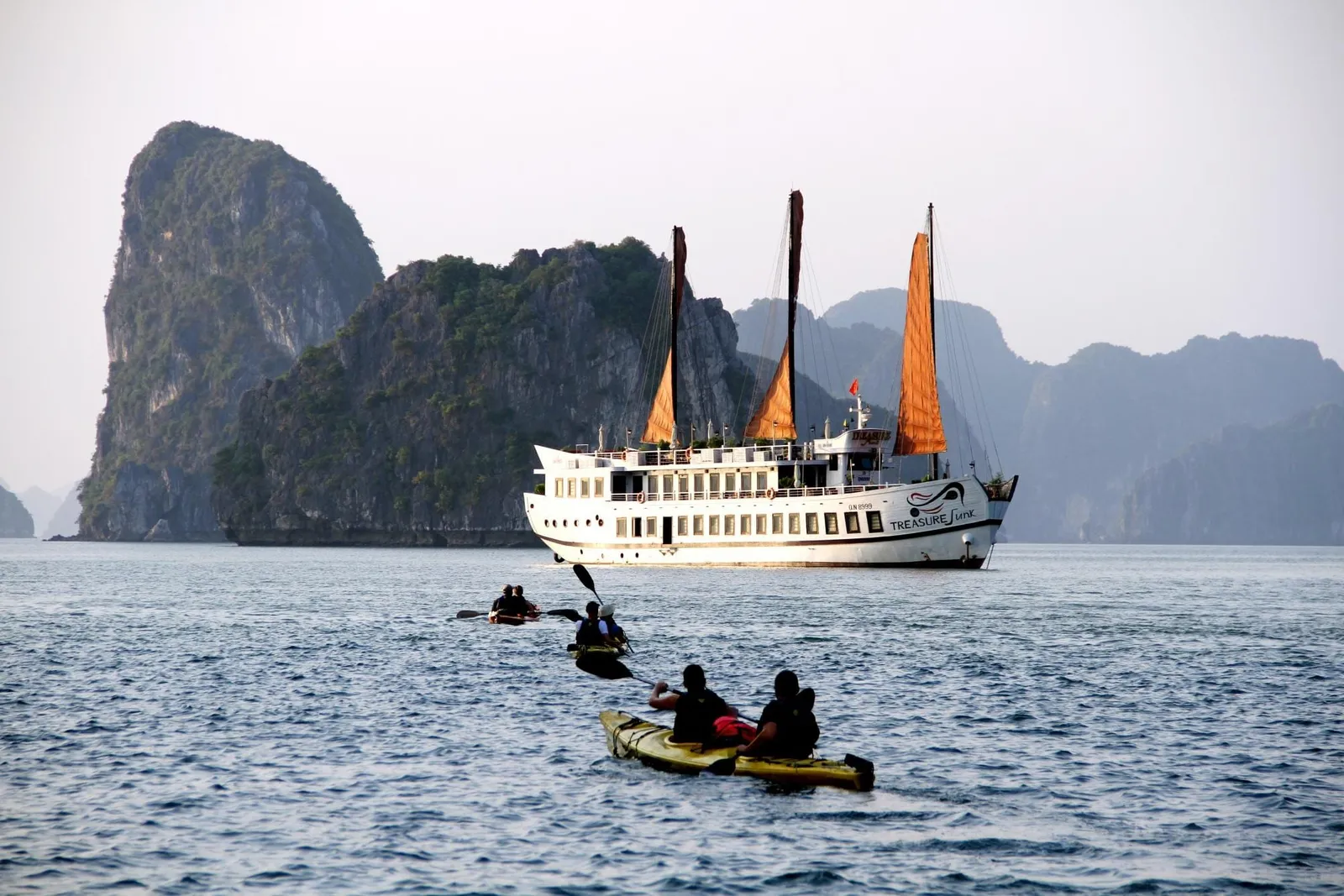 A white three-masted cruise boat with orange sails and 'Treasure Junk' lettering on its hull sits in front of limestone karst cliffs while kayakers paddle in the foreground.