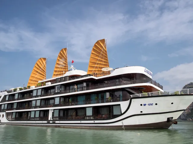A multi-deck white cruise ship with three orange junk-style sails and 'La Regina' lettering on its hull and sails floats on green water beside a limestone karst under a blue sky with clouds.