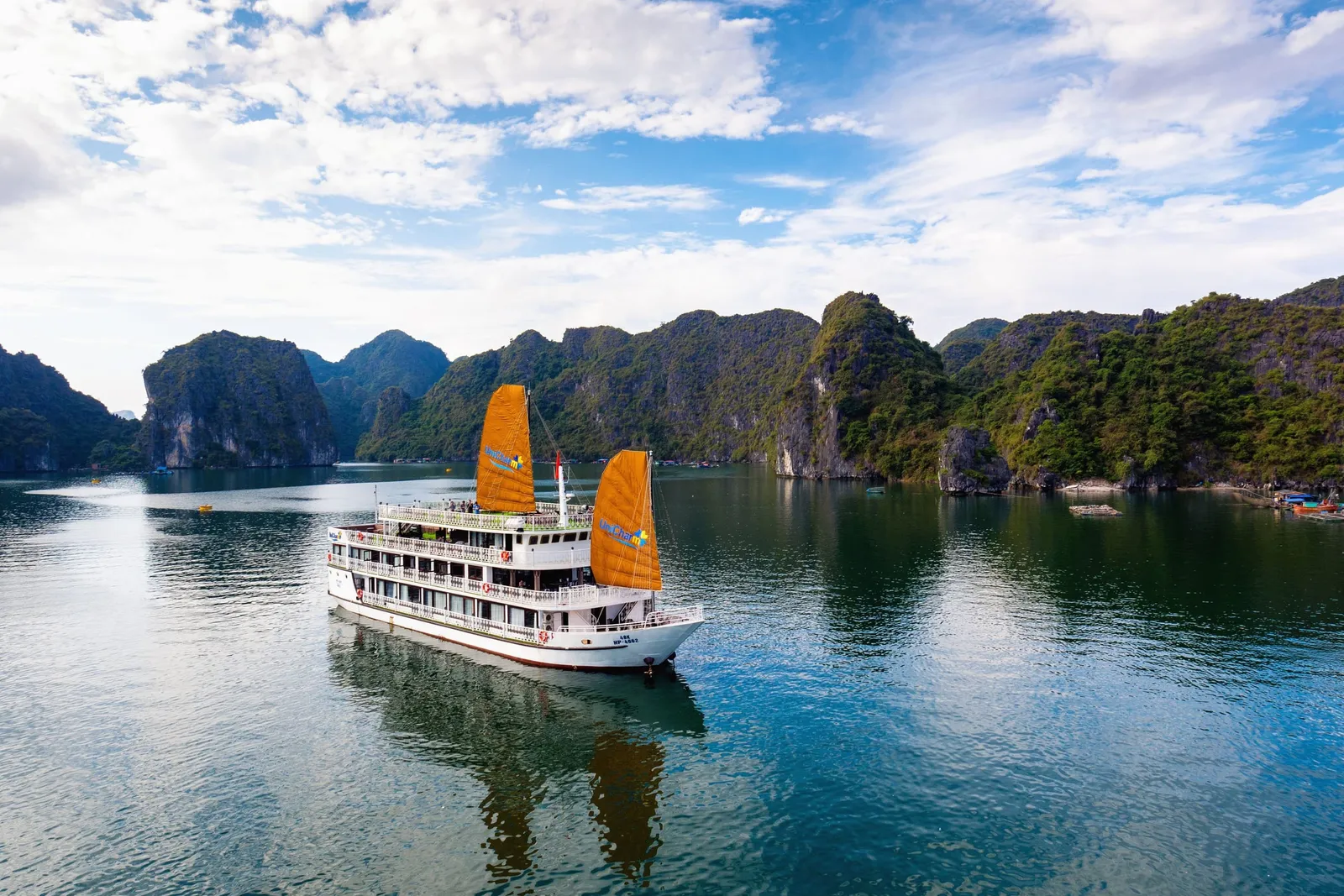 A white multi-deck cruise boat with two orange sails and logos on the sails and hull floats on calm green water in front of forested limestone karsts under a partly cloudy blue sky, photographed from an elevated, drone-like angle.