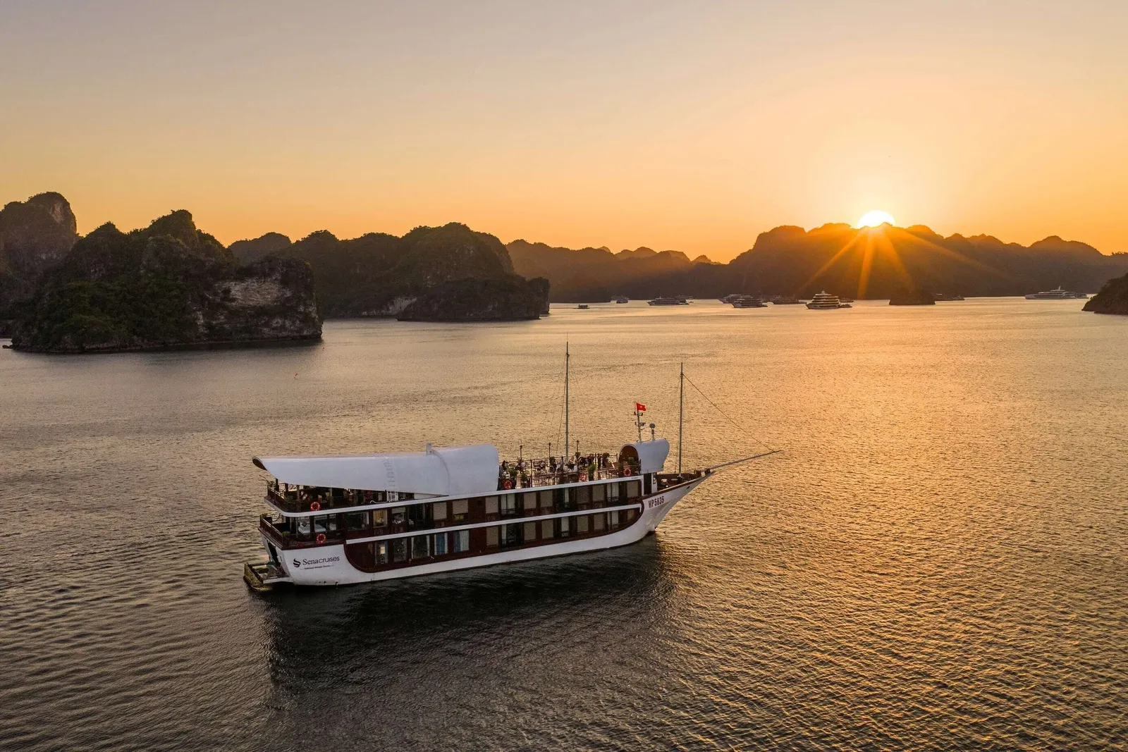 An aerial view of a white and dark-red multi-deck cruise boat with a sail-like canopy and a company logo on its hull, sitting on calm water at sunset among limestone karst islands with other boats in the distance.