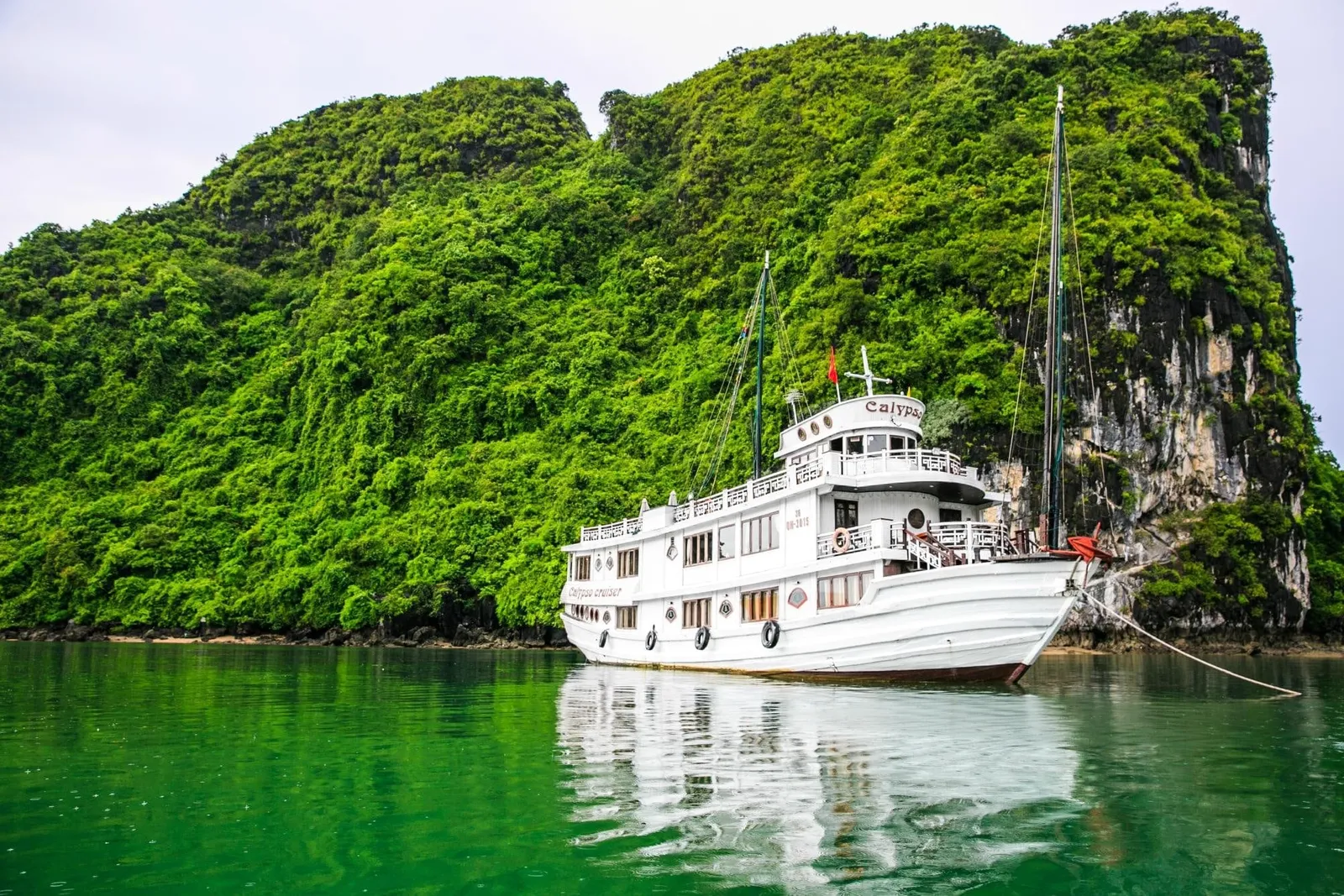 A white two-deck vessel with bare masts and no sails sits moored on green water in front of a steep, forested limestone island.