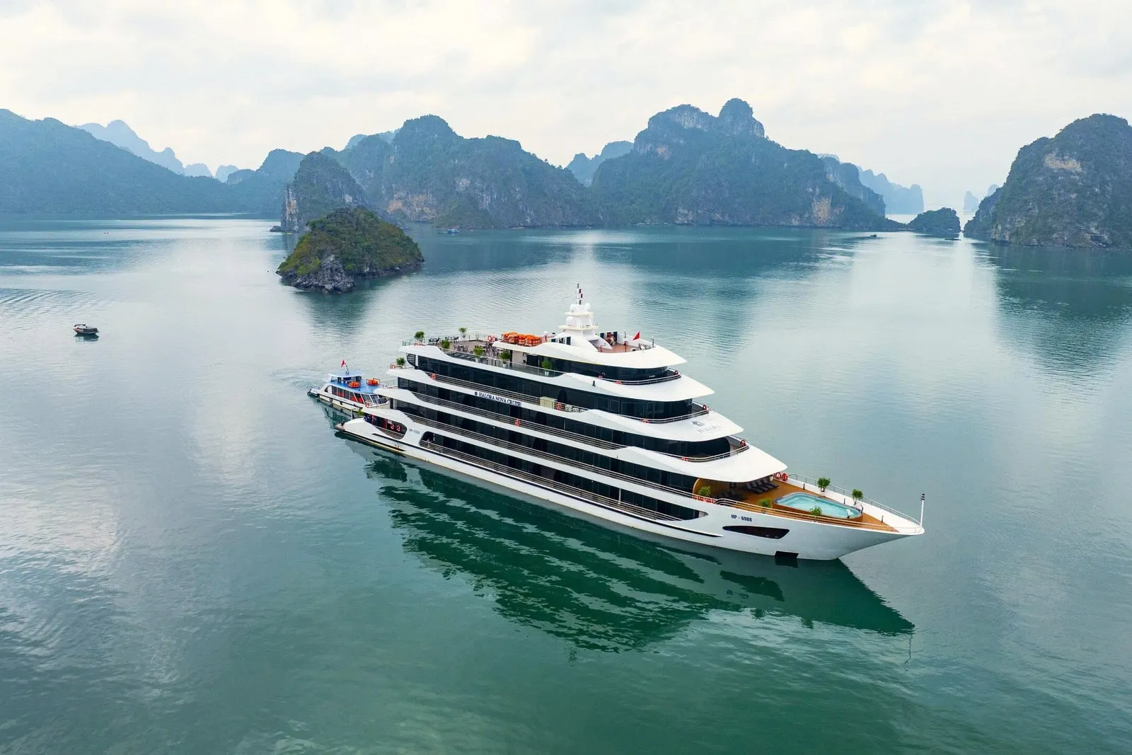 An aerial view of a large white multi-deck cruise ship with a small pool at the stern, sitting on calm green water among limestone karst islands, with a small boat in the distance and small lettering on the hull.