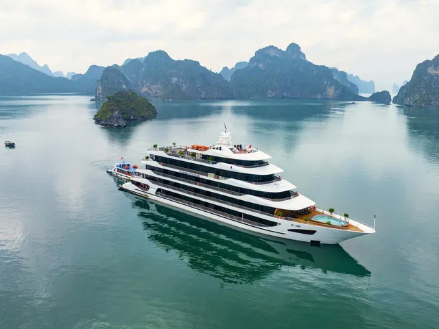 An aerial view of a large white multi-deck cruise ship with a small pool at the stern, sitting on calm green water among limestone karst islands, with a small boat in the distance and small lettering on the hull.