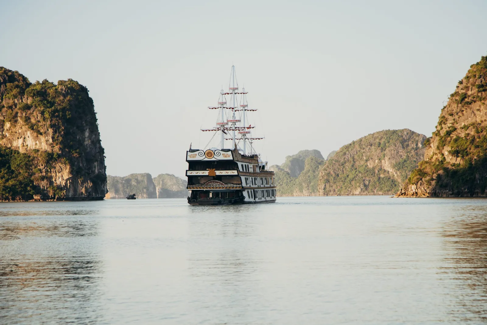 A multi-deck black and white cruise ship with tall masts and decorative stern sits on calm water between green limestone karst islands under a pale sky.