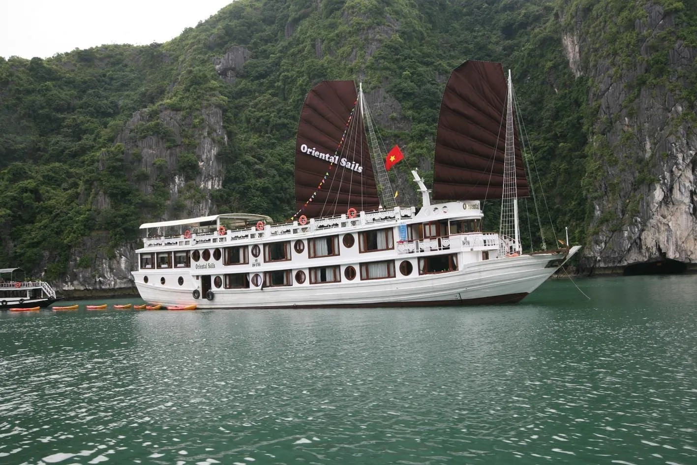 A white cruise junk with dark brown sails reading 'Oriental Sails' and a Vietnamese flag sits on green water in front of steep tree-covered limestone cliffs, with kayaks lined up beside it and another boat at left.