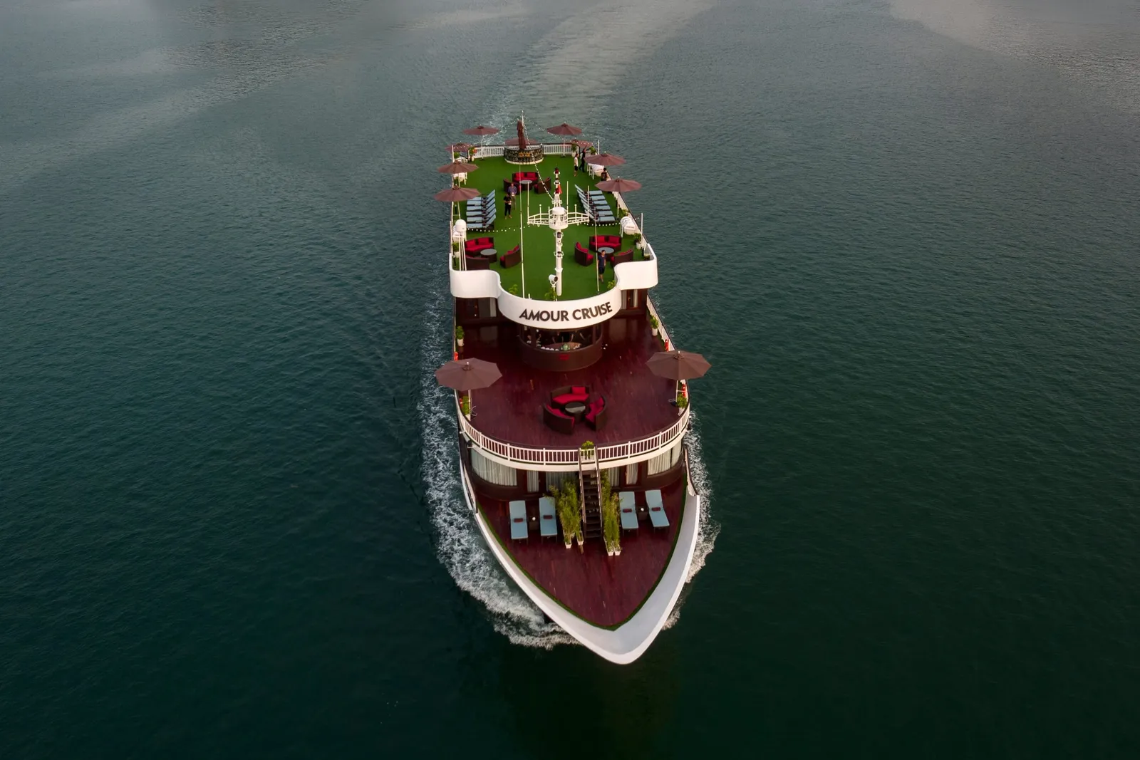 Aerial view of a multi-deck cruise boat with 'AMOUR CRUISE' lettered on its upper deck facade, moving through dark green water with a white wake, with umbrellas, lounge seating and sun loungers on its decks.