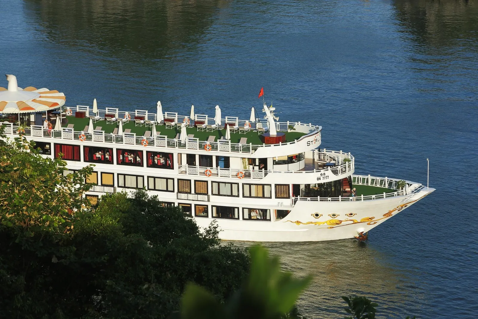 A white multi-deck cruise boat with a golden dragon design on the bow, a registration number (QN 7777) and a partly legible name on the hull, seen from a high vantage point above dark green foliage on calm blue water.