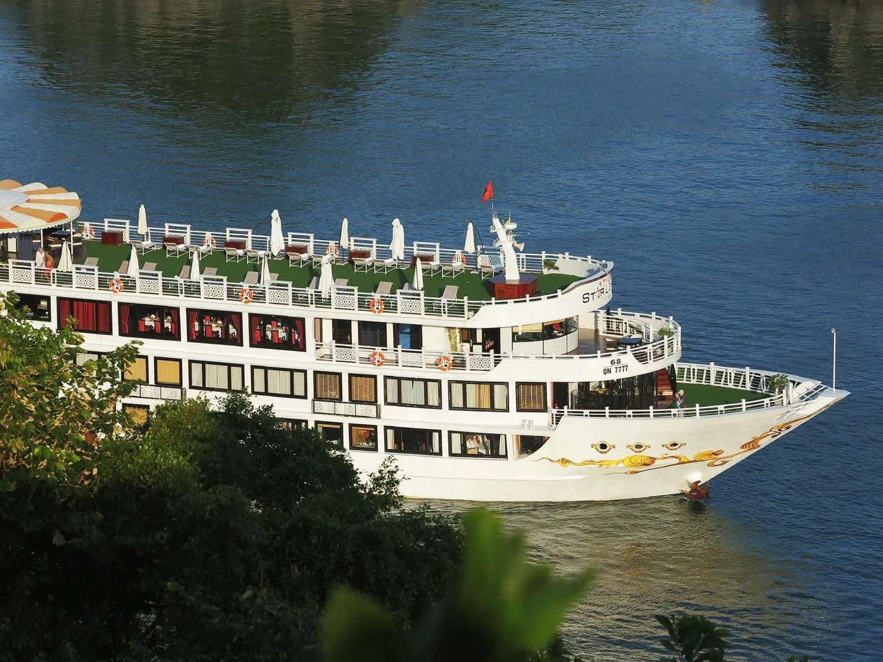 A white multi-deck cruise boat with a golden dragon design on the bow, a registration number (QN 7777) and a partly legible name on the hull, seen from a high vantage point above dark green foliage on calm blue water.