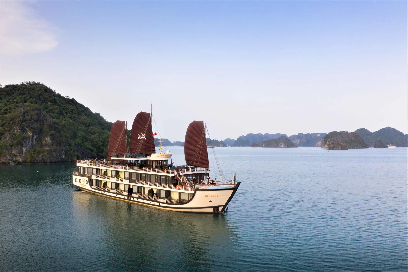 A multi-deck white cruise boat with three dark red sails bearing a white lotus-style emblem sits on calm green water in front of forested limestone islands, photographed from an elevated outside viewpoint.