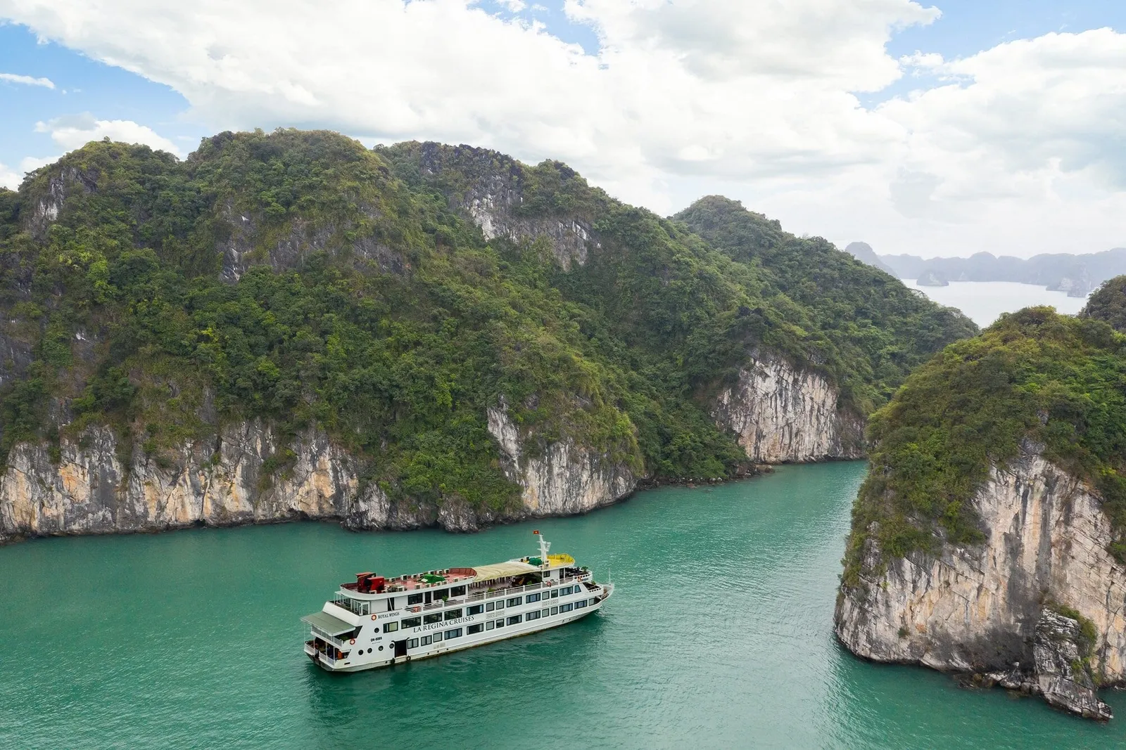 Aerial view of a white multi-deck cruise ship with 'La Regina Cruises' lettering on its hull sitting on emerald water between forested limestone karst cliffs under a cloudy blue sky.