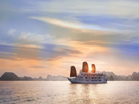 A white multi-deck cruise boat with three dark orange sails, some with a small emblem, floats on calm water at sunset with limestone karst silhouettes behind it.