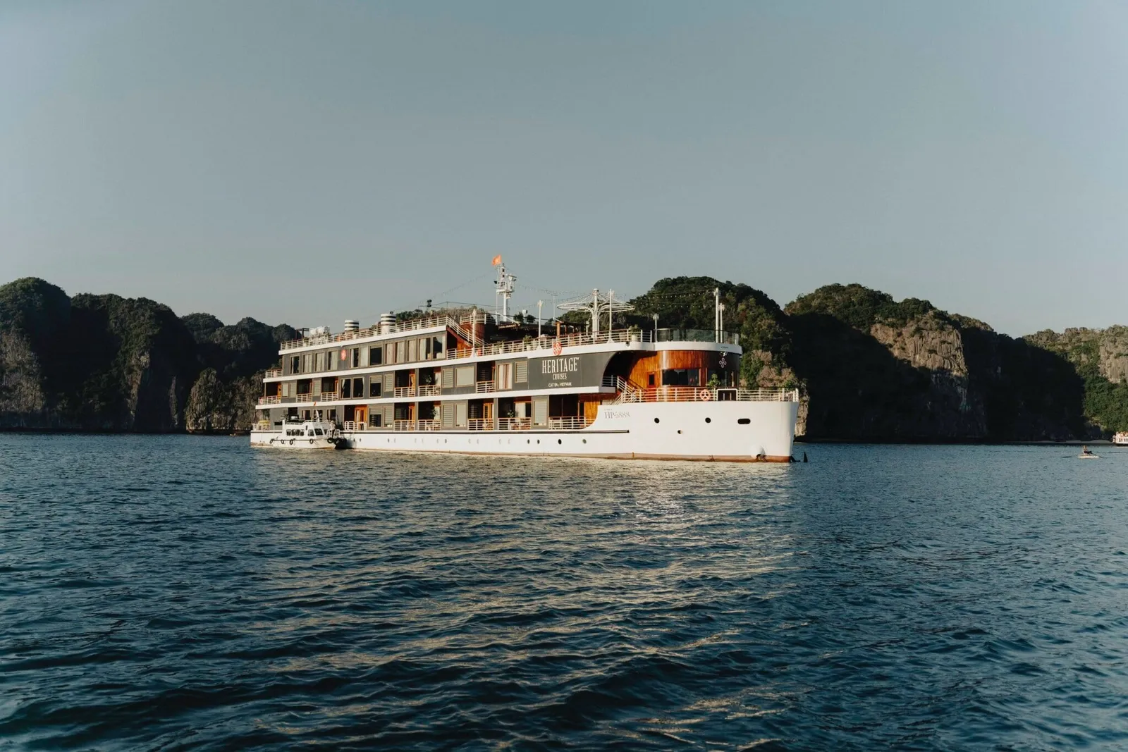 A multi-deck white cruise ship with 'Heritage Cruises' lettering on its side sits on calm blue water in front of tree-covered limestone karst islands under a clear sky.
