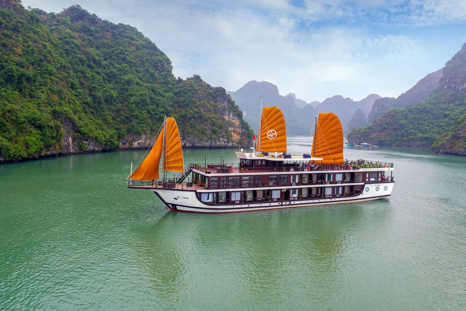 A multi-deck cruise boat with orange sails, a flower-shaped logo on one sail and small text on the hull, floats on green water between forested limestone karst hills under a hazy sky, photographed from above and outside the vessel.