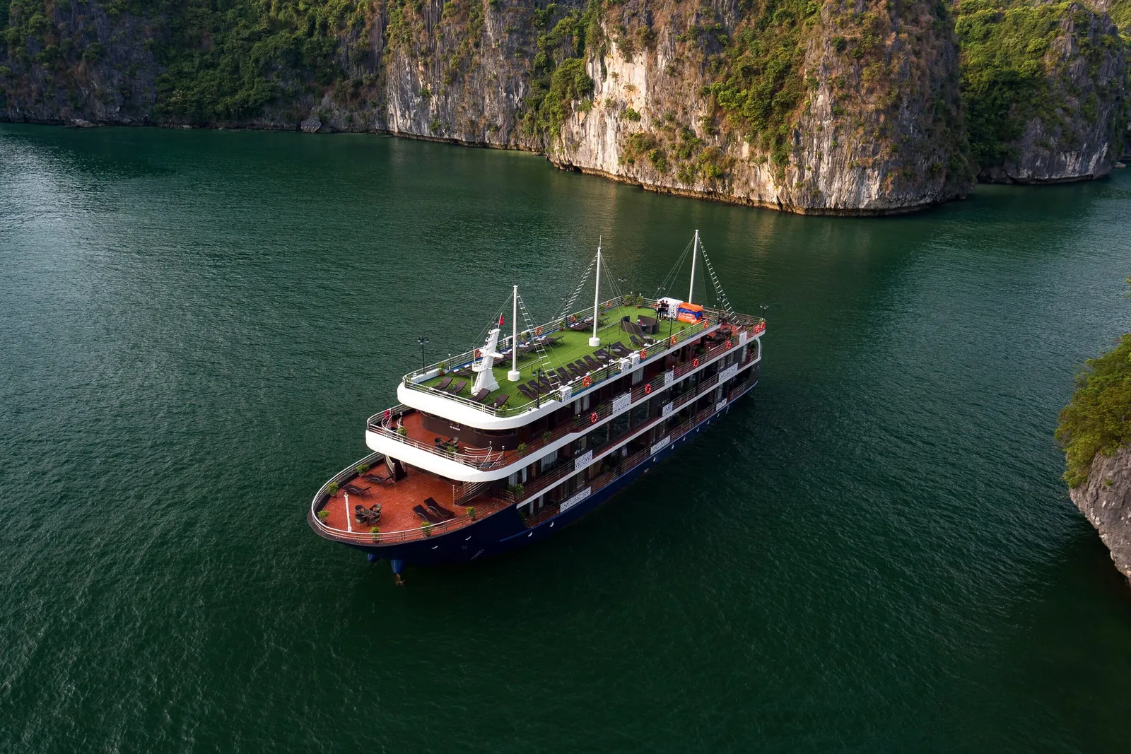 Aerial view of a three-deck cruise ship with a navy hull, wooden lower decks and a green artificial-turf top deck, anchored on dark green water beside steep limestone cliffs.