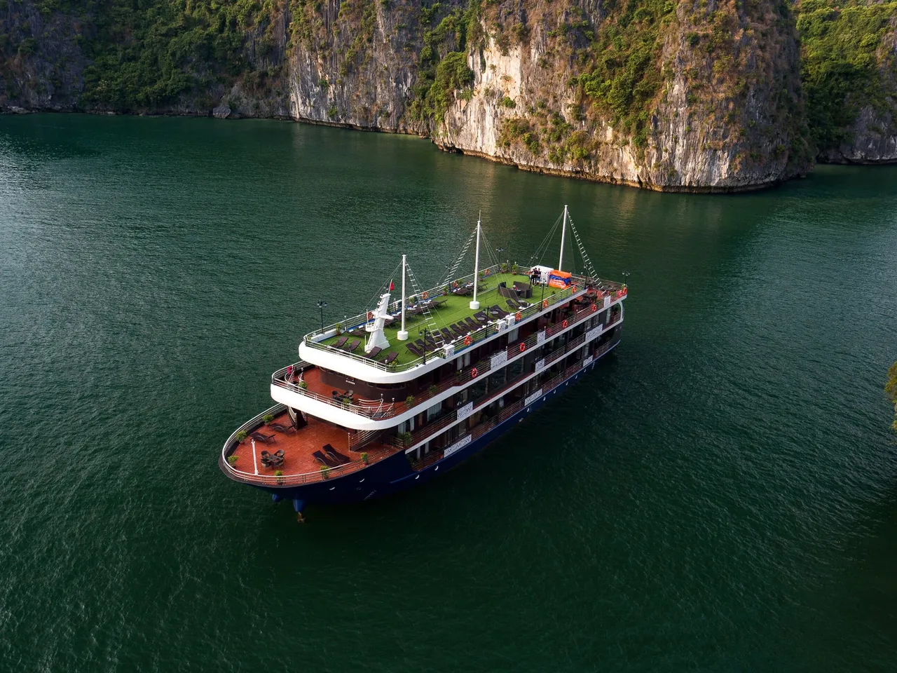 Aerial view of a three-deck cruise ship with a navy hull, wooden lower decks and a green artificial-turf top deck, anchored on dark green water beside steep limestone cliffs.