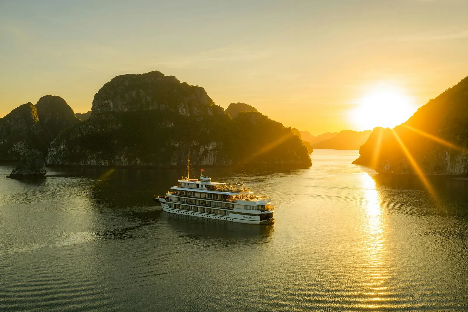 An aerial view of a multi-deck white cruise ship on calm water among karst limestone islands at sunset, with the sun low on the right and a small name painted on the stern.