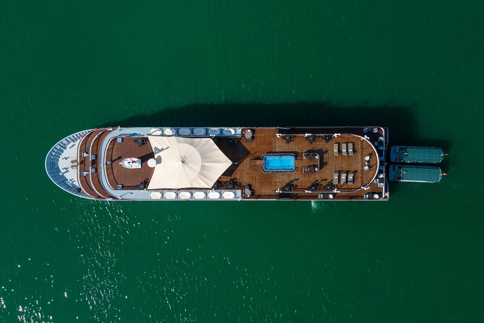 Top-down aerial view of a cruise ship on green water, showing a wooden sun deck with a small pool, a cream canopy, loungers, and two tender boats at the stern.
