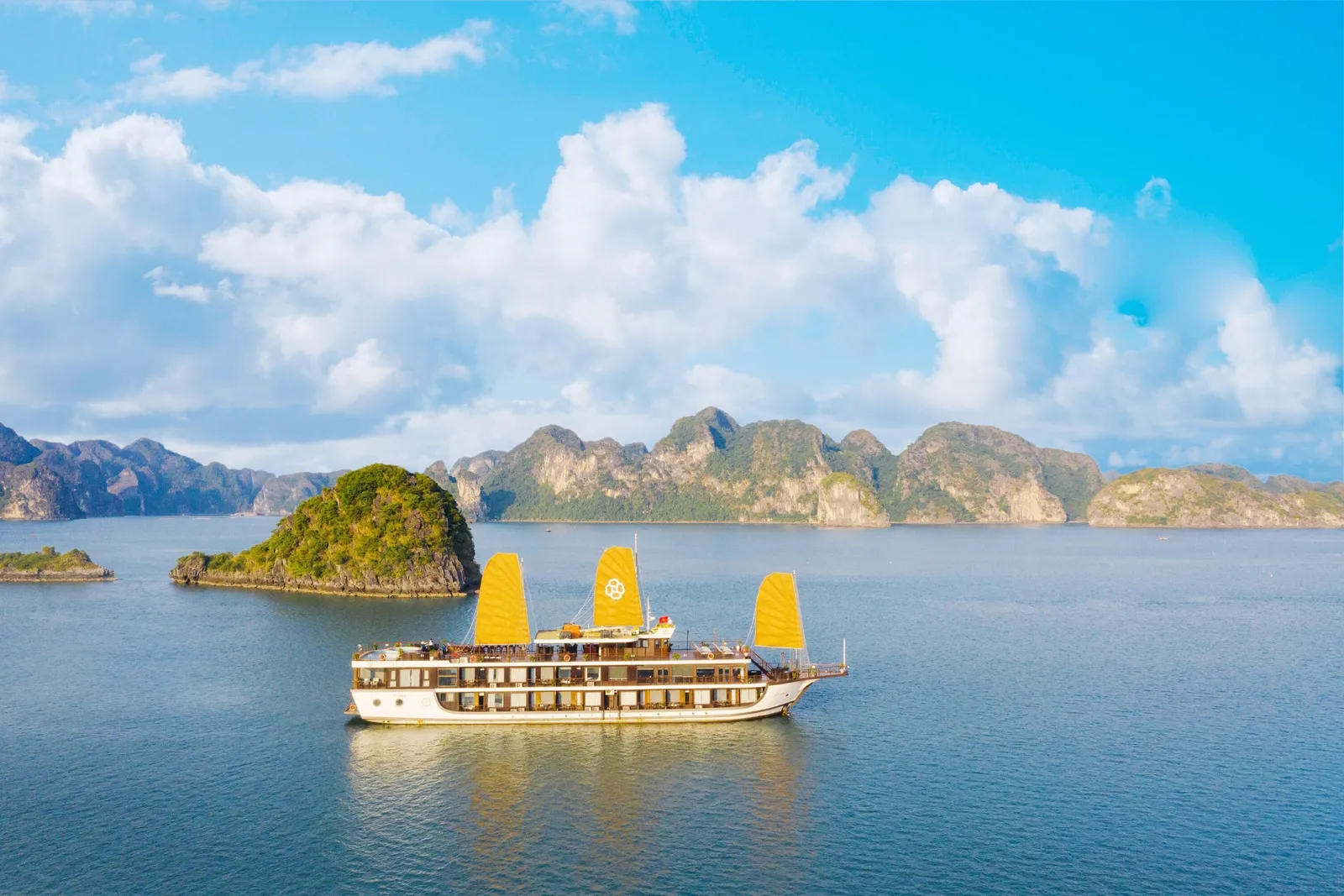 A white multi-deck cruise ship with three yellow junk-style sails, one bearing a circular emblem, floats on calm blue water in front of green limestone karst islands under a blue sky with large clouds, seen from an elevated outside viewpoint.