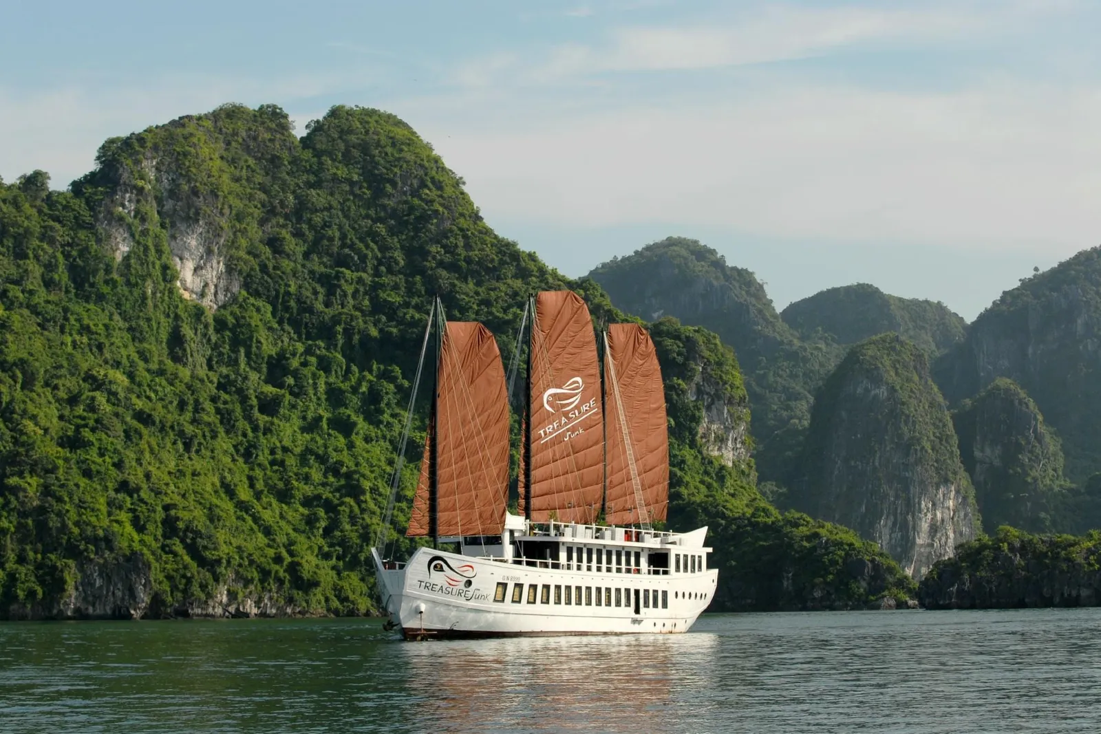 A white junk-style boat with three rust-brown sails and 'Treasure Junk' branding on the hull and middle sail floats on calm water in front of green limestone karst hills.