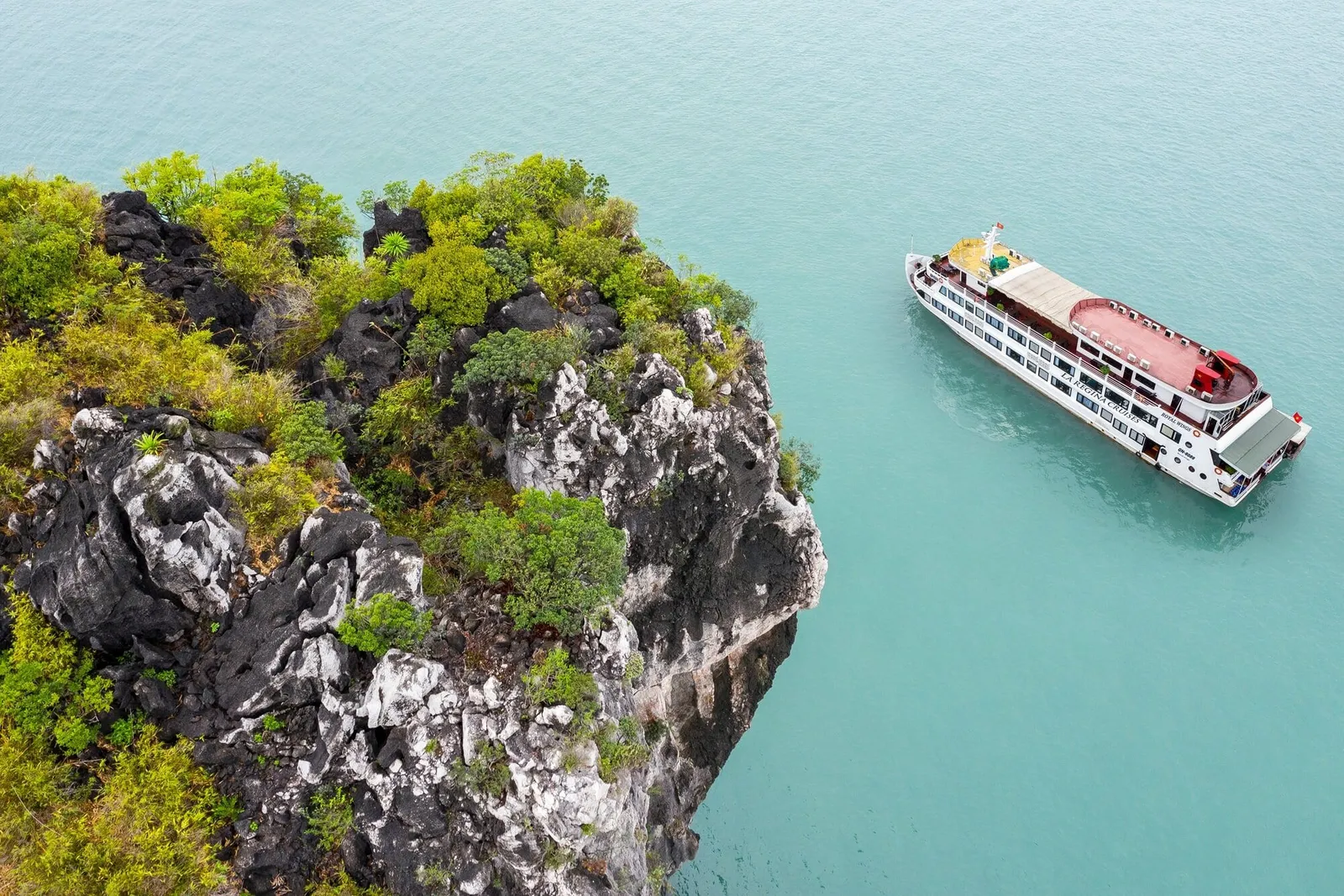 An aerial view of a white multi-deck cruise ship with a red-topped upper deck anchored in turquoise water beside a rocky, vegetation-covered limestone outcrop, with faint lettering on the ship's hull.