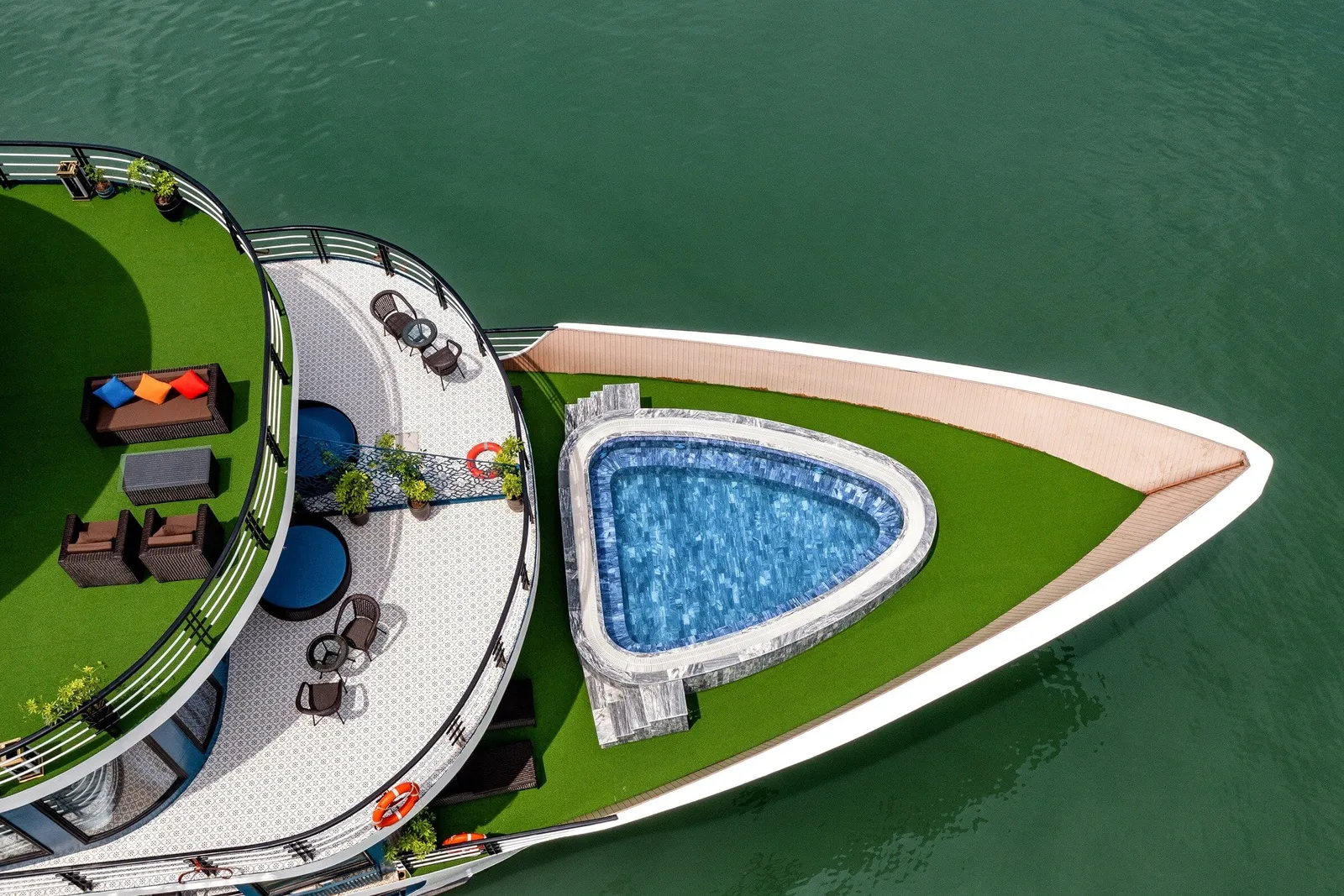Aerial top-down view of a cruise ship bow with a triangular blue-tiled pool on artificial grass, upper decks with wicker seating, cushions, potted plants and life rings, surrounded by green water.