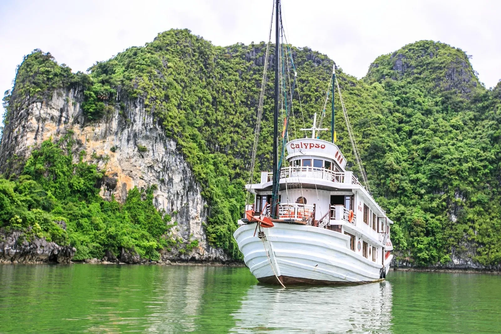 A white two-deck wooden cruise boat named Calypso, anchored on green water in front of steep limestone karst covered in vegetation.
