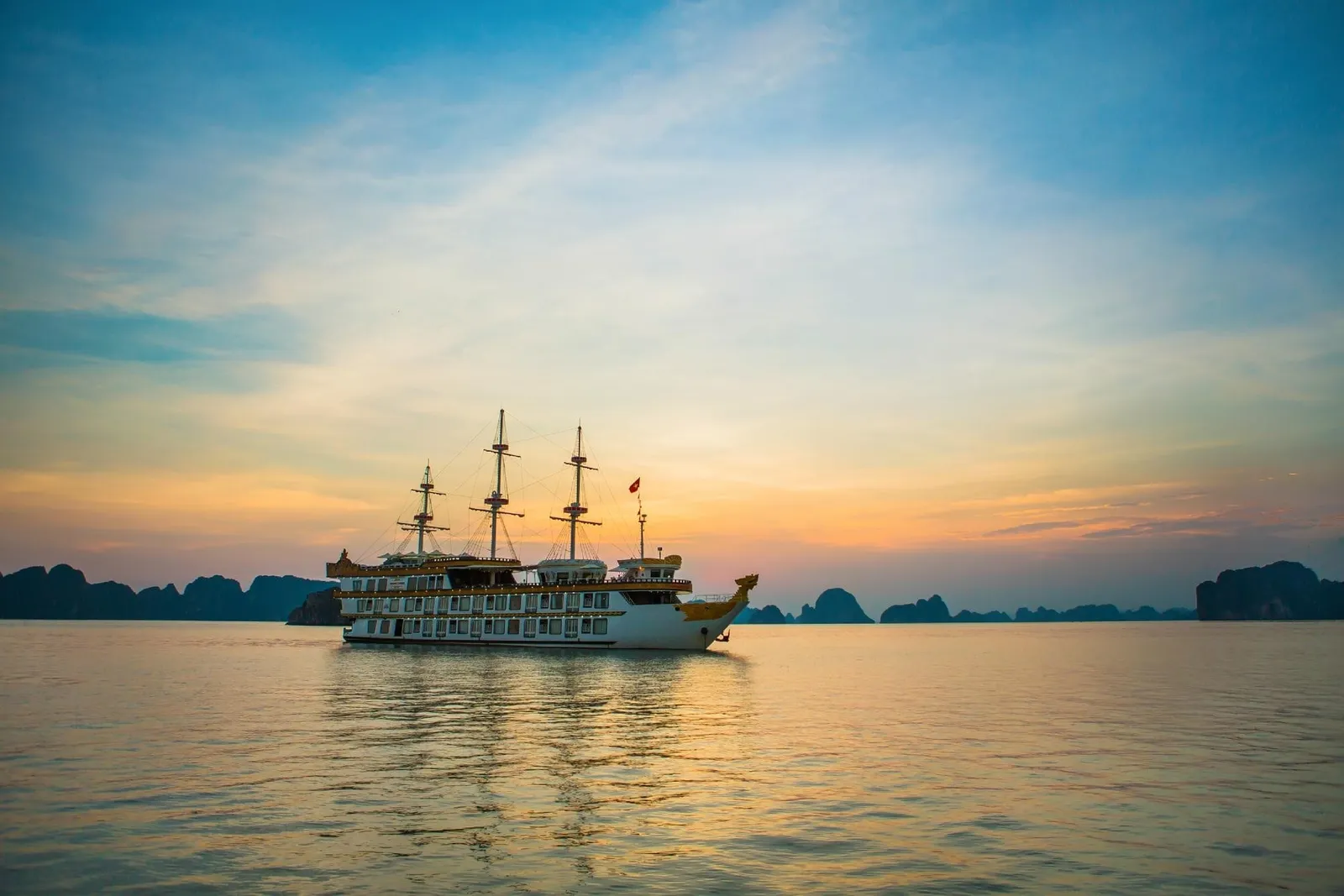 A three-masted multi-deck cruise ship with a golden dragon-style bow floats on calm water at sunset, with dark limestone karst silhouettes on the horizon.