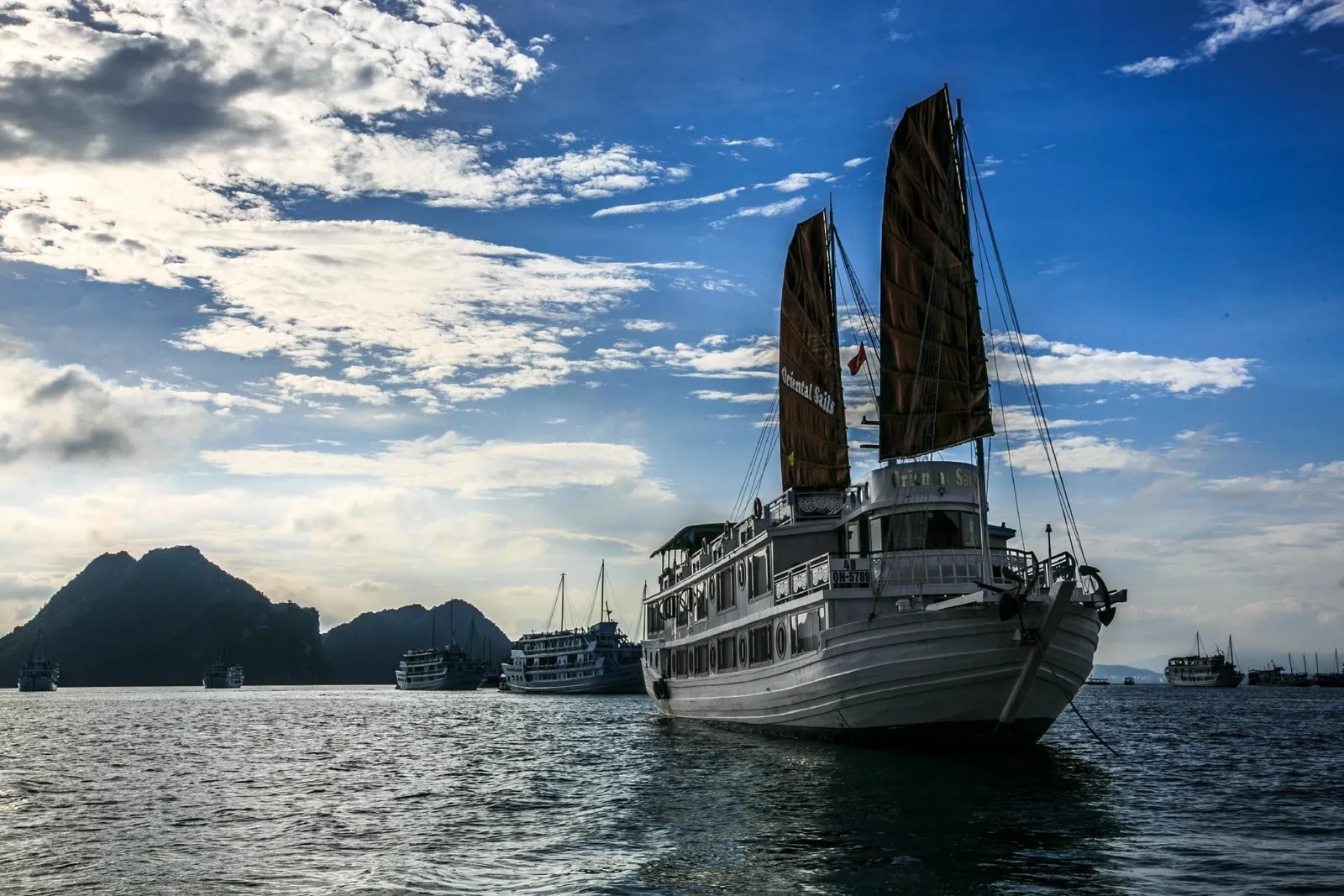 A white two-deck wooden junk with dark brown sails sits at anchor on the bay at dusk, with limestone karsts and other boats in the background.