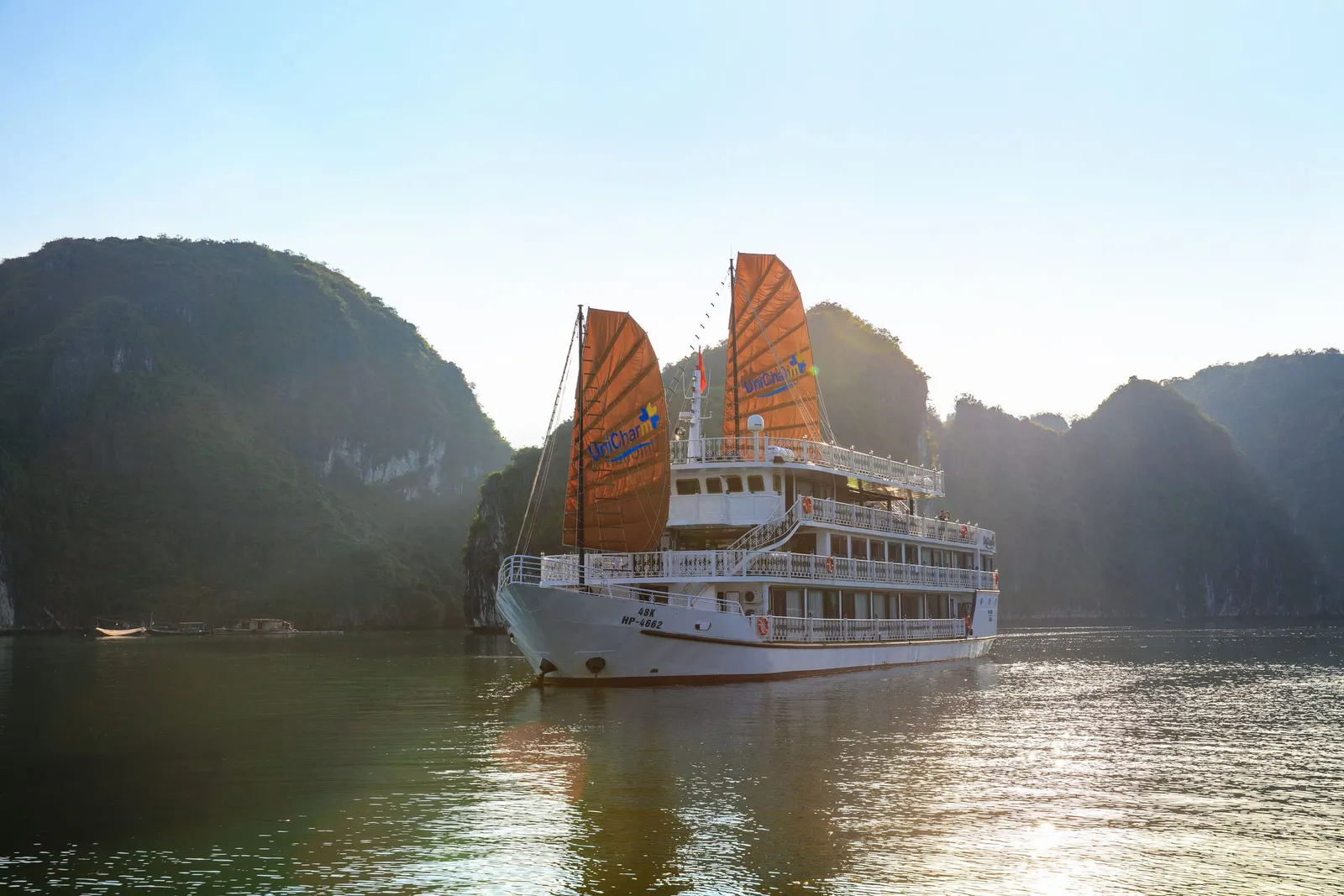 A white multi-deck cruise boat with two orange sails bearing a blue logo sits on calm green water in front of forested limestone karst hills, with hull registration text visible and hazy sunlit sky above.