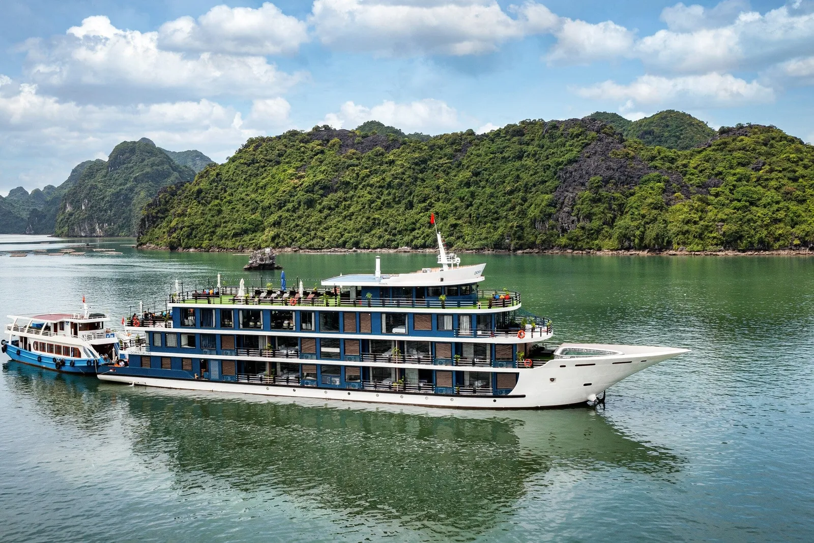 A large white and blue multi-deck cruise ship on green water with a smaller blue and white tender boat alongside, in front of forested limestone karst islands under a partly cloudy sky.