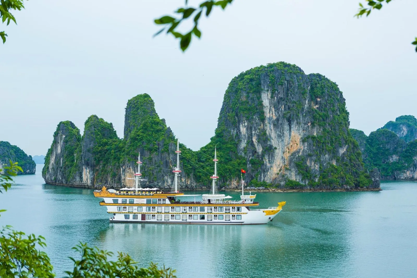 A white and gold multi-deck three-masted cruise ship with a Vietnamese flag sails on calm green water in front of tall limestone karst islands, framed by leaves in the foreground.