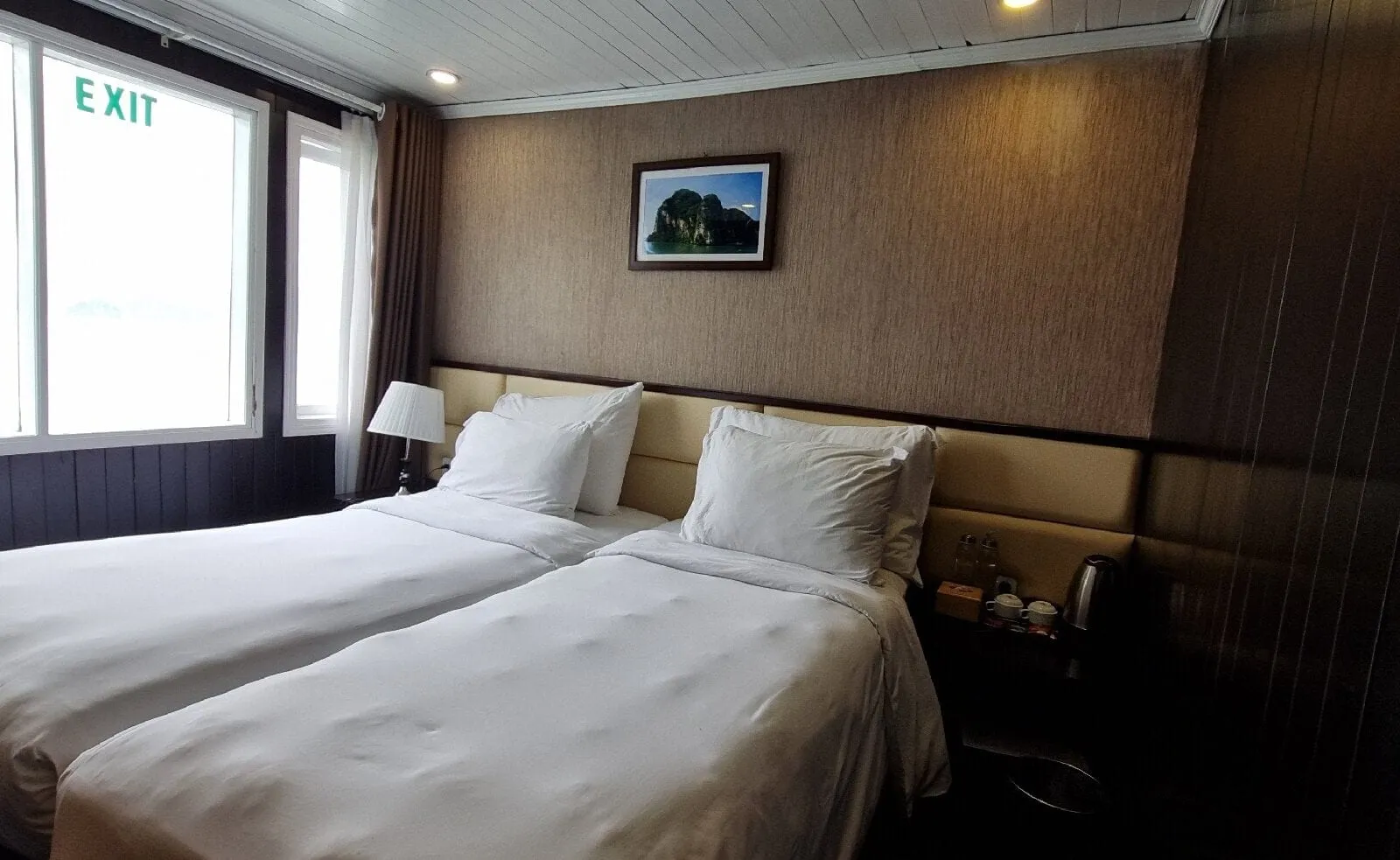 A cabin with two twin beds pushed together with white linens, a framed limestone karst picture on the wall, a window with a green EXIT sign, and a wall-mounted air conditioner remote.