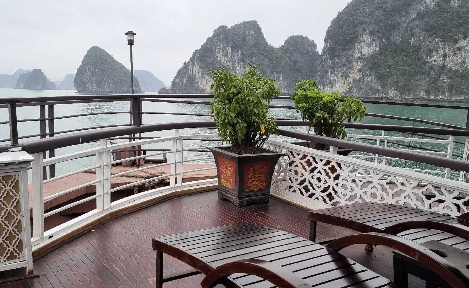 A wooden upper deck with white lattice railings, loungers and potted plants overlooks calm green water and misty limestone karst islands under an overcast sky.